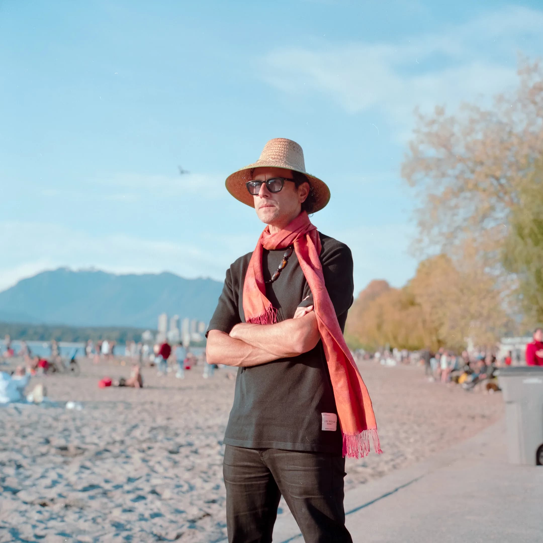 Clemente Castillo standing on a sandy beach with arms crossed, wearing a straw hat, sunglasses, and a coral-pink scarf with mountains in the background